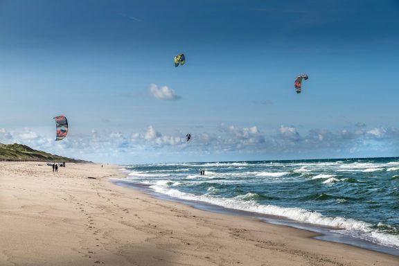 Nordsee & Surffeeling Kiter gleiten über das Wasser vor einem weitläufigen Strand unter blauen Himmel.